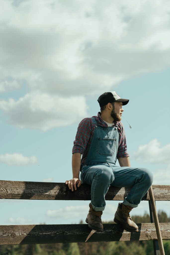 A farmer in denim overalls and cap sits on a fence, gazing into the distance in the countryside.