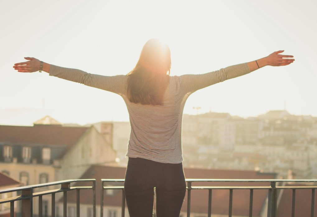 A woman stands with outstretched arms on a sunny balcony, embracing the morning light.