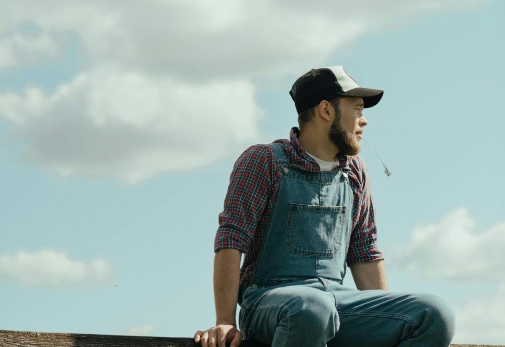 A farmer in denim overalls and cap sits on a fence, gazing into the distance in the countryside.